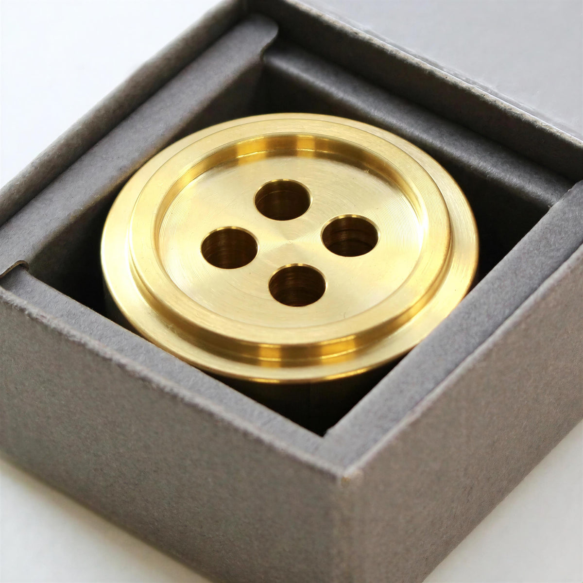 A close-up image of a golden button-like object, possibly a decorative piece or a small sculpture, placed inside a gray box.
