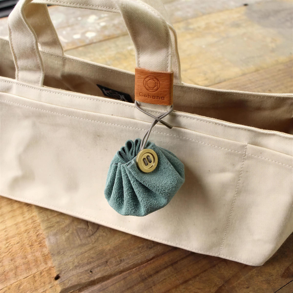 A beige canvas tote bag with a small, round, green coin purse attached to it, resting on a wooden surface.