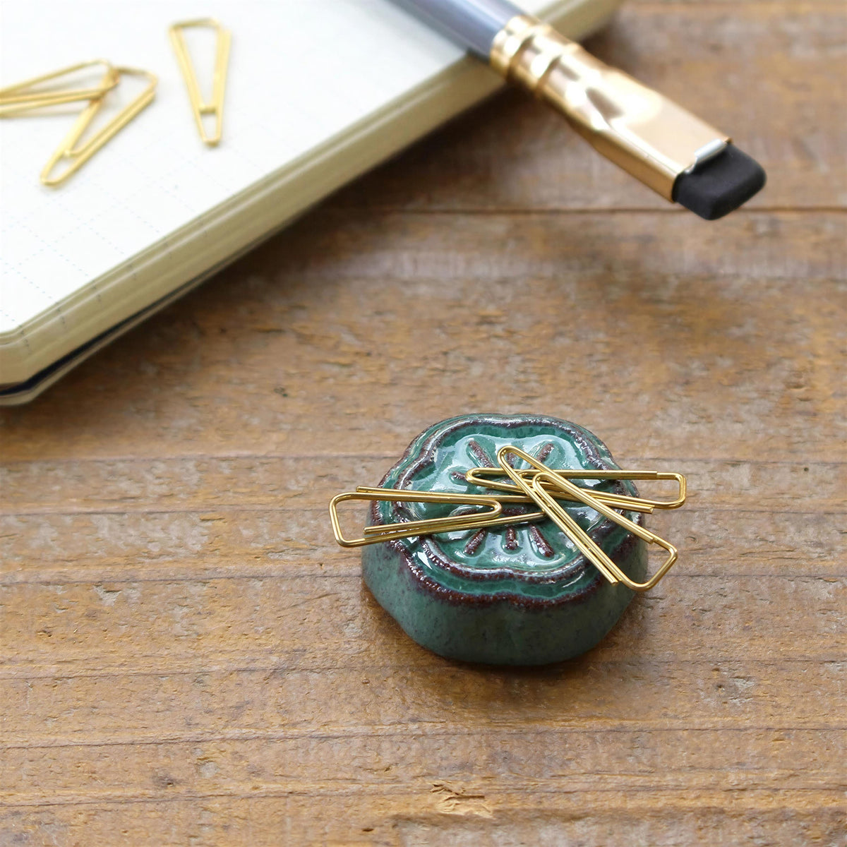a close-up view of a green ceramic object with gold-colored metal clips attached to it, placed on a wooden surface alongside a pen and a notebook.