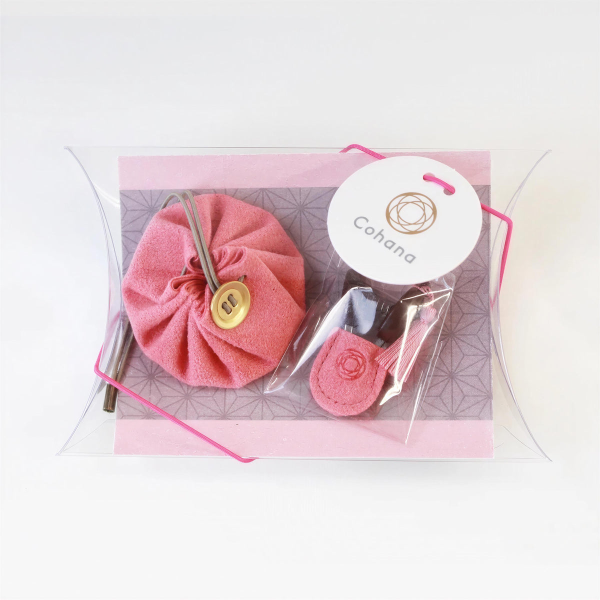 A pink pouch with a gold button, a pink keychain, and a pink tag with the word "Cohana" printed on it, all placed in a clear plastic container with a pink ribbon.