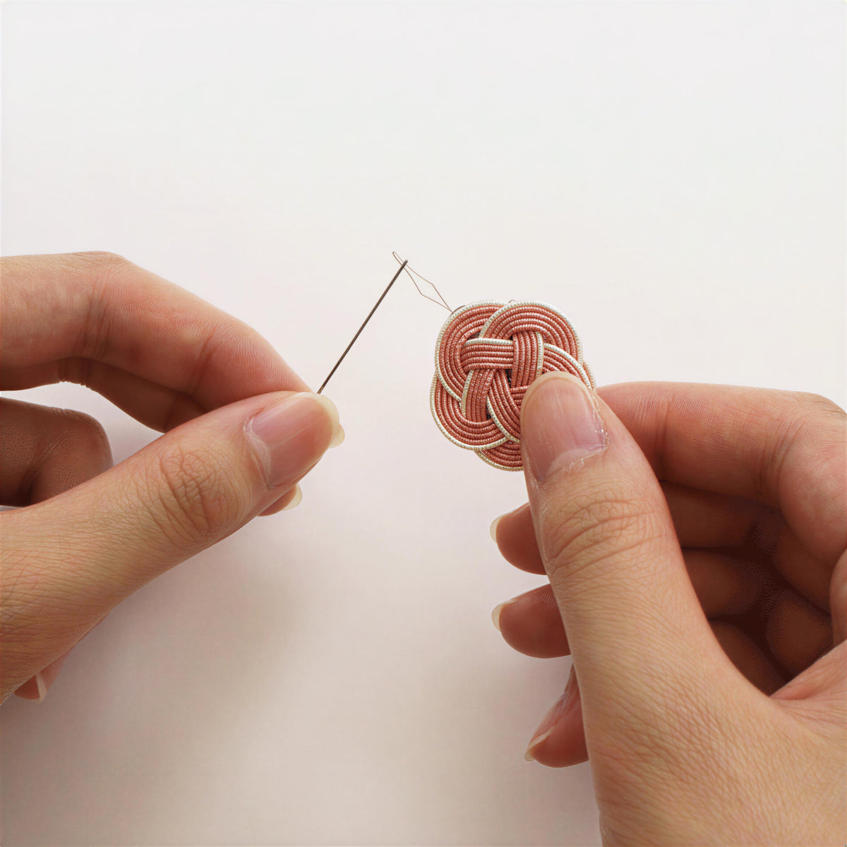 A pair of hands holding a needle and thread, with the needle piercing through a small, intricately woven object.