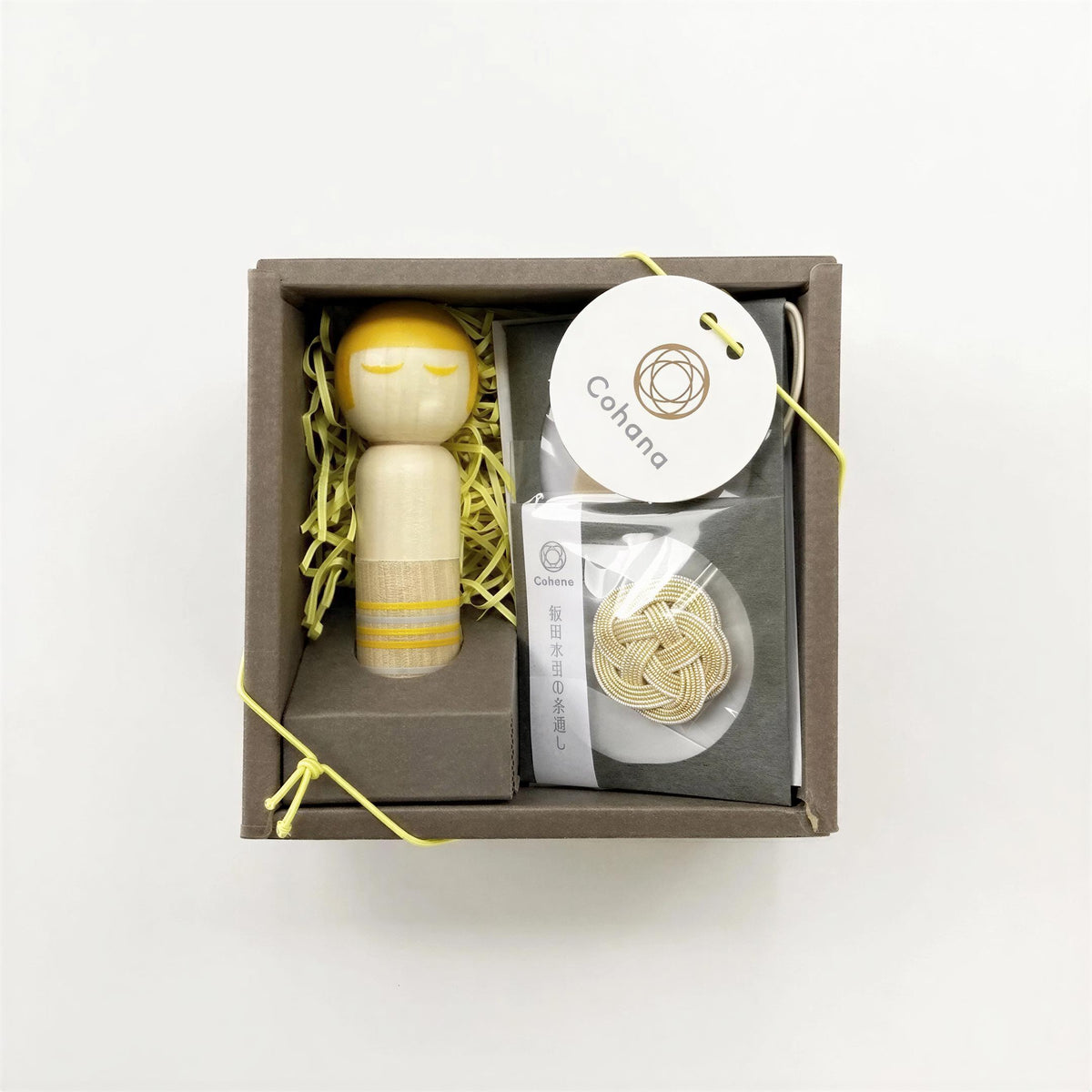 A small wooden figurine with a yellow head and arms is displayed in a brown box, accompanied by a white label and a small package of string.