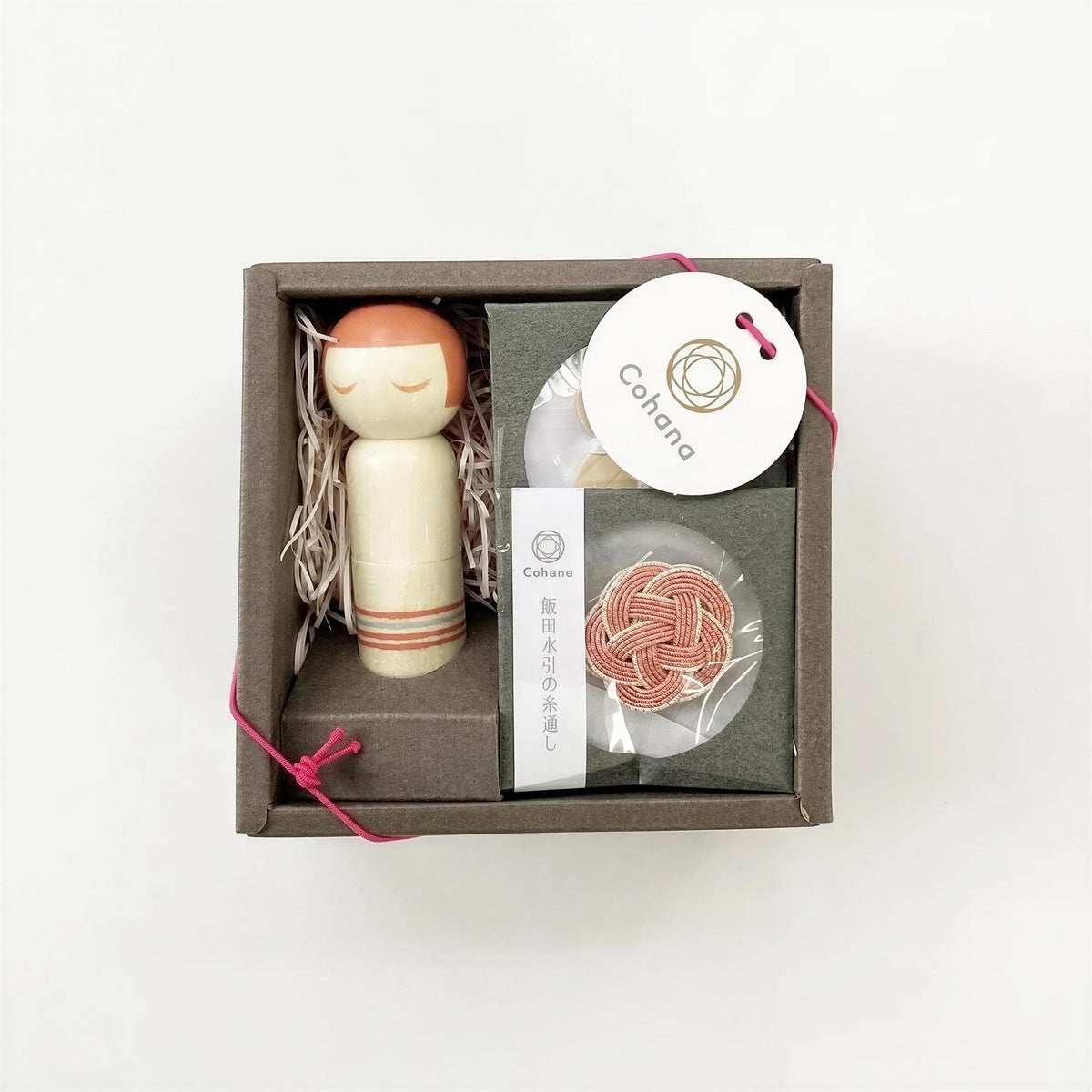 A small wooden doll with a pink head and a white body, along with some string and a small white box with a label, all placed in a brown box with a pink ribbon.
