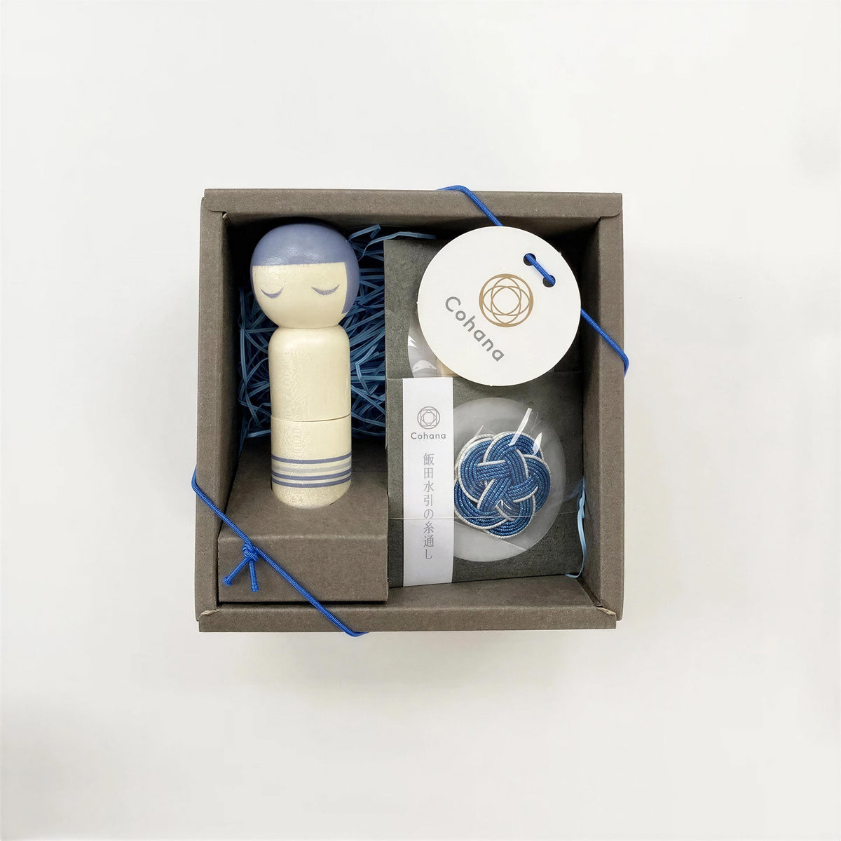 A small wooden doll with blue eyes and a blue ribbon, along with some other items, all placed in a brown box with a white label.