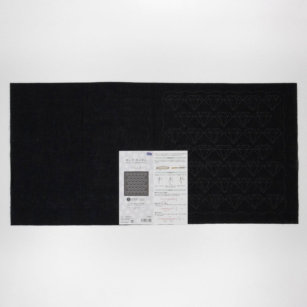 The image is a black and white photograph of a large piece of fabric with a diamond pattern on it, along with a small white label or instructions booklet placed next to it.