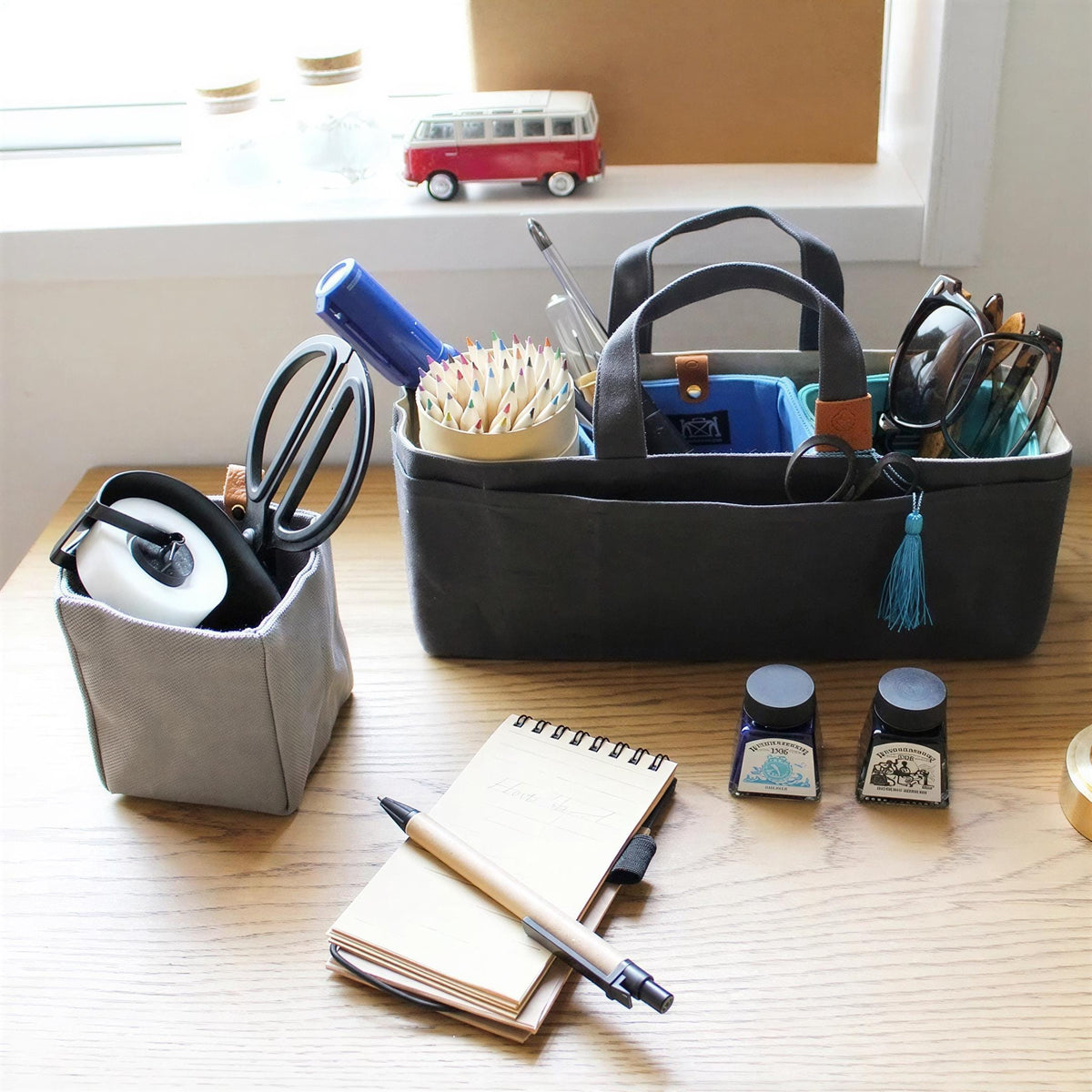 a wooden desk with various items on it, including a gray container holding a pair of scissors, a notebook, a pen, and a small toy bus.