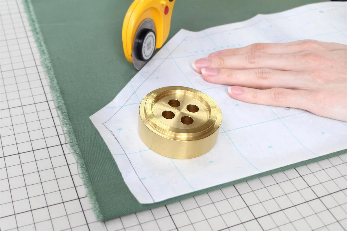A hand is cutting a piece of fabric with a yellow and black scissors, while a brass button is placed on a piece of paper with a grid pattern.
