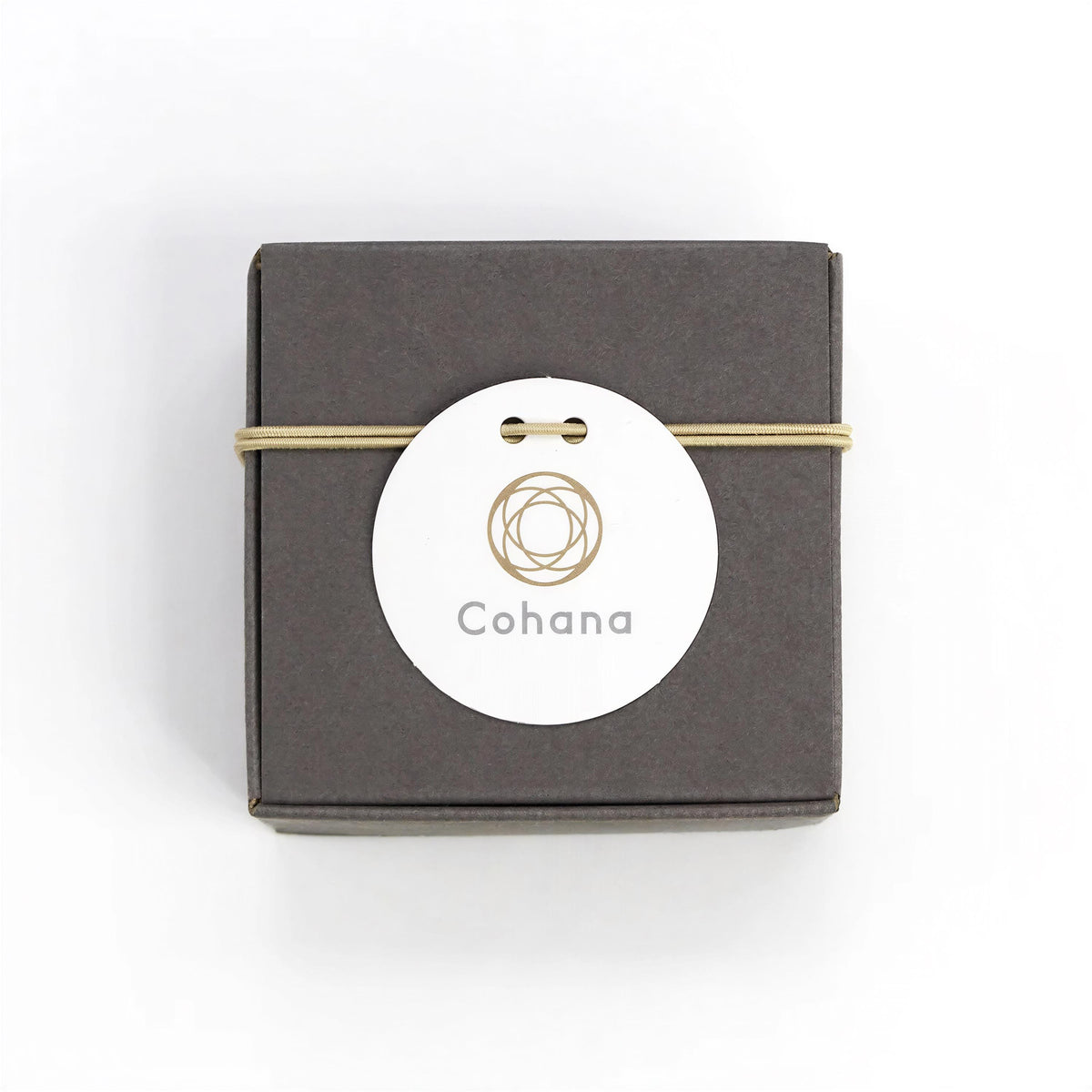 A square-shaped dark grey box with a white circular label featuring a gold design and the word "Cohana" printed on it.