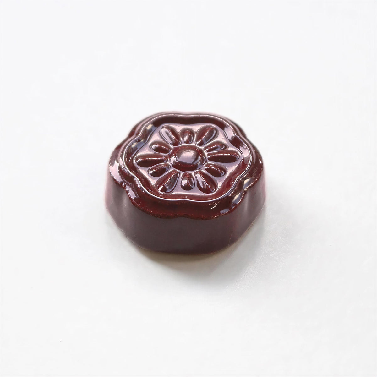 A single, shiny, round, dark brown object with a flower-like design on top, sitting on a white surface.