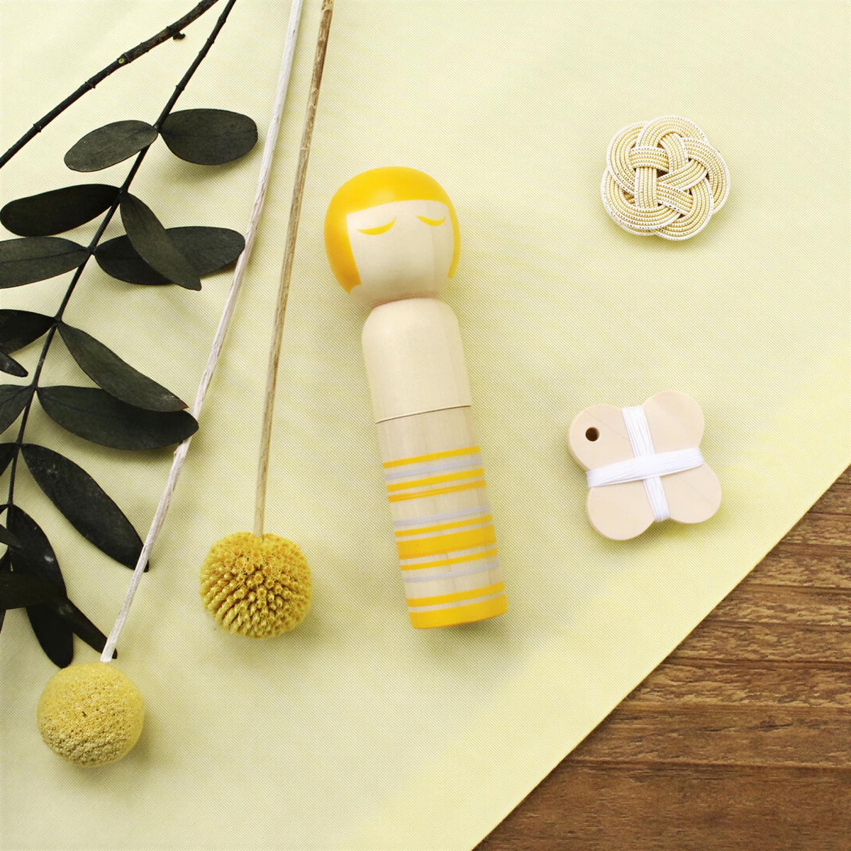 a wooden doll with a yellow head and body, a small wooden bead, and some dried leaves and flowers on a yellow surface.