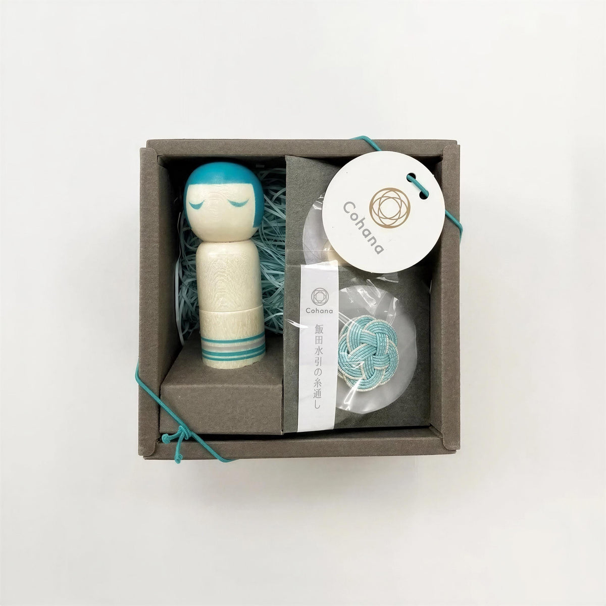 A small wooden doll with a teal head and a white body, placed inside a gray box with a white label and a blue string.