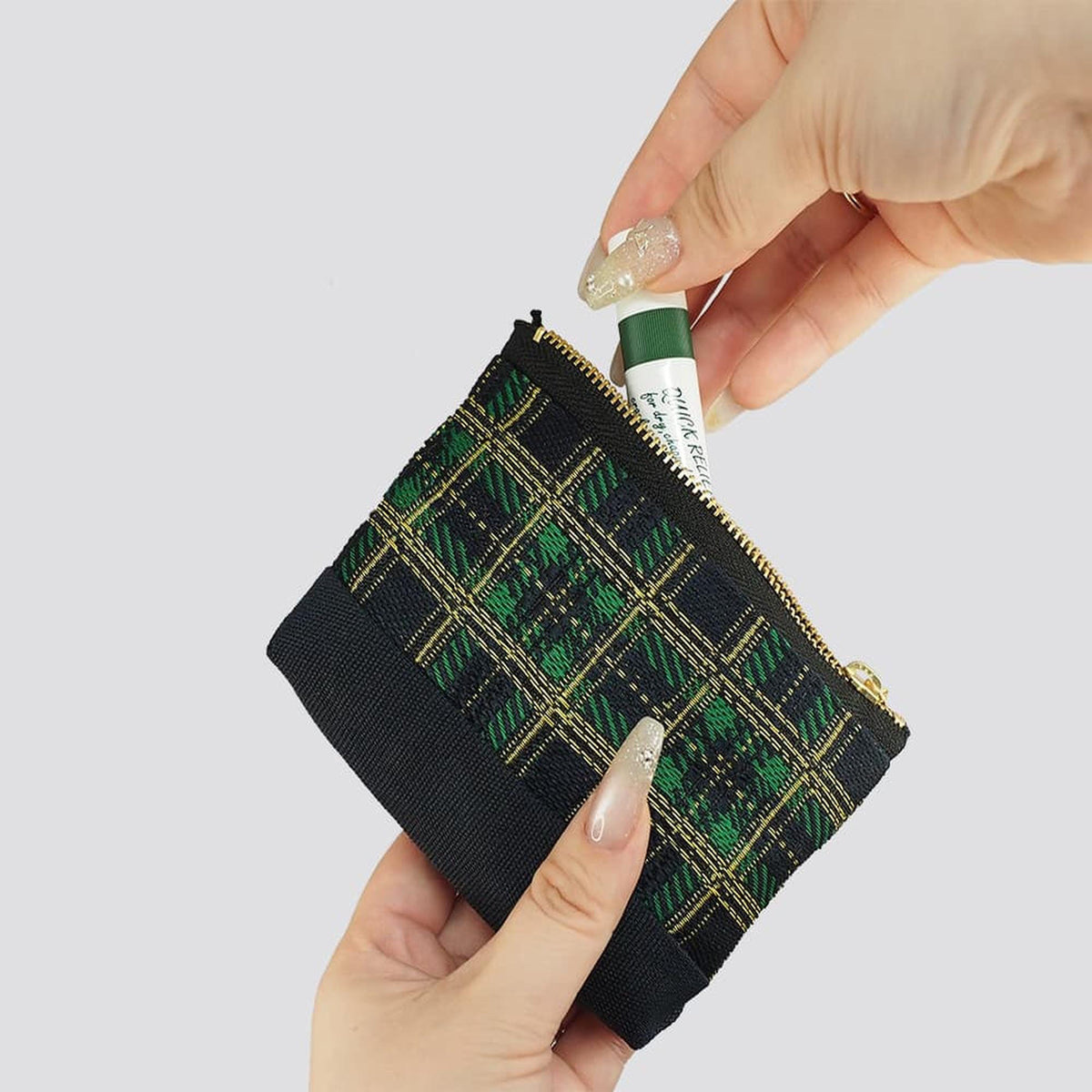 A hand holding a black and green zippered pouch with a green and gold pattern, and a tube of lip balm inside the pouch.