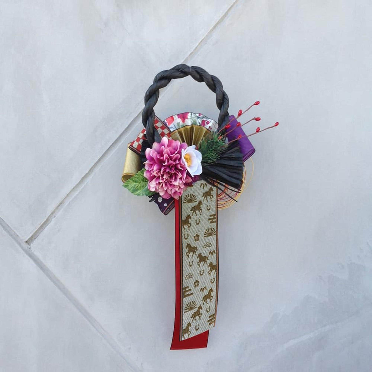 A decorative wreath made of various materials, including a black braided handle, a pink flower, and a gold and red patterned ribbon.