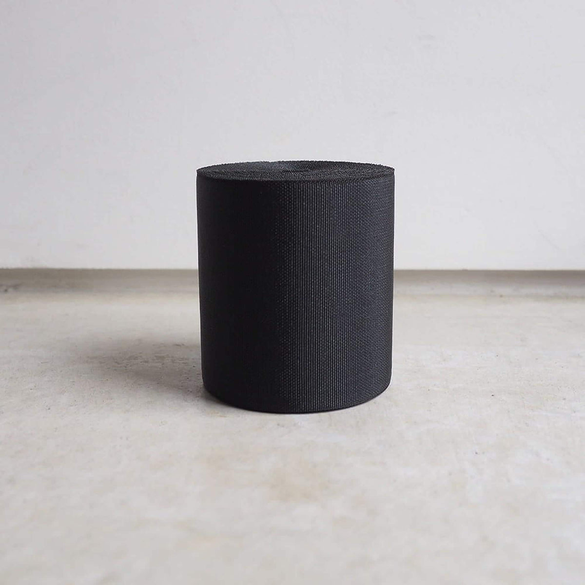 A black cylindrical object is standing alone on a concrete floor against a plain white wall.