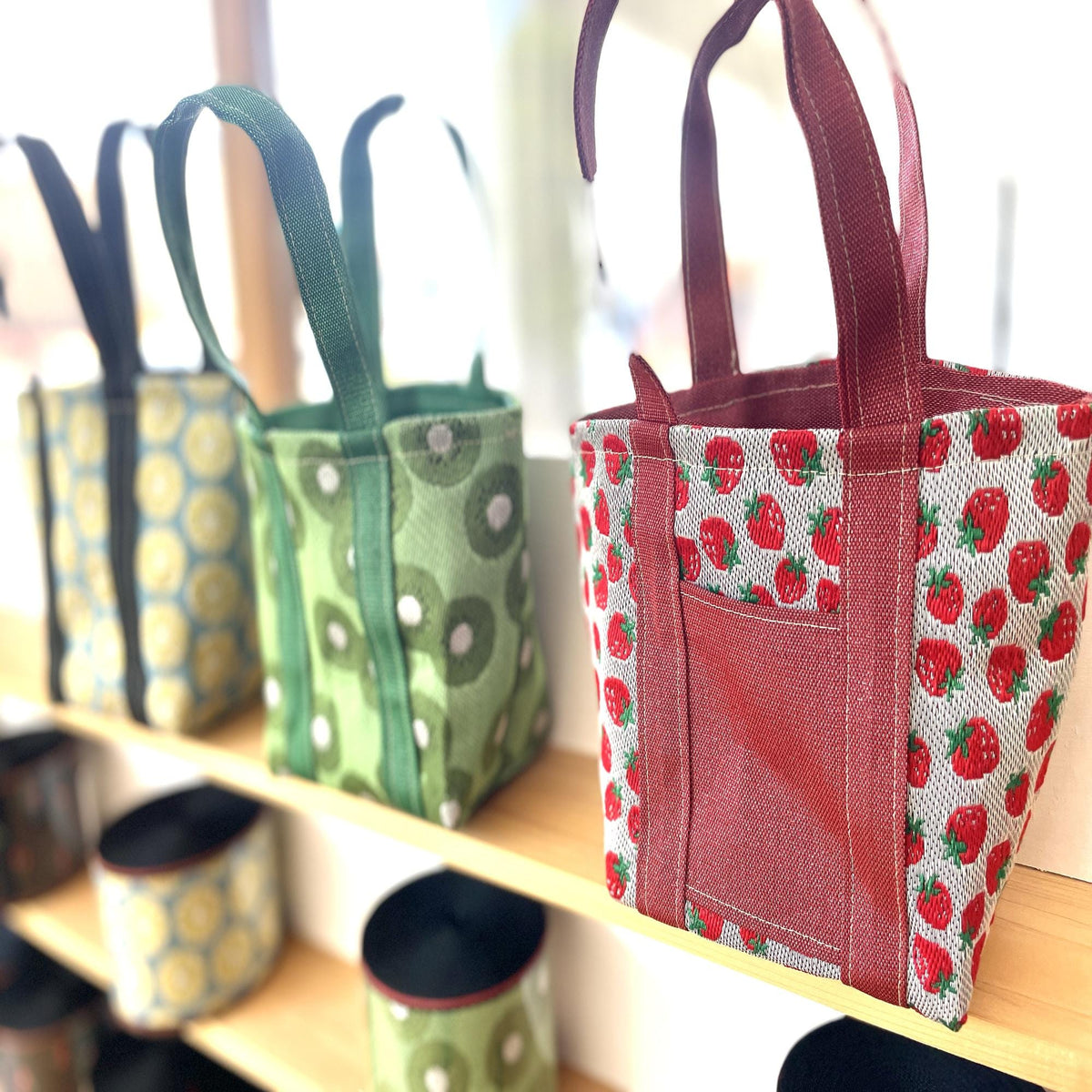 A row of colorful tote bags with various patterns, including green, red, and white designs, displayed on a shelf.
