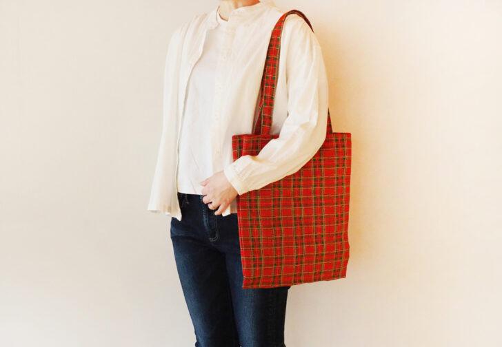 A person wearing a white shirt and blue jeans is holding a red and green plaid tote bag.