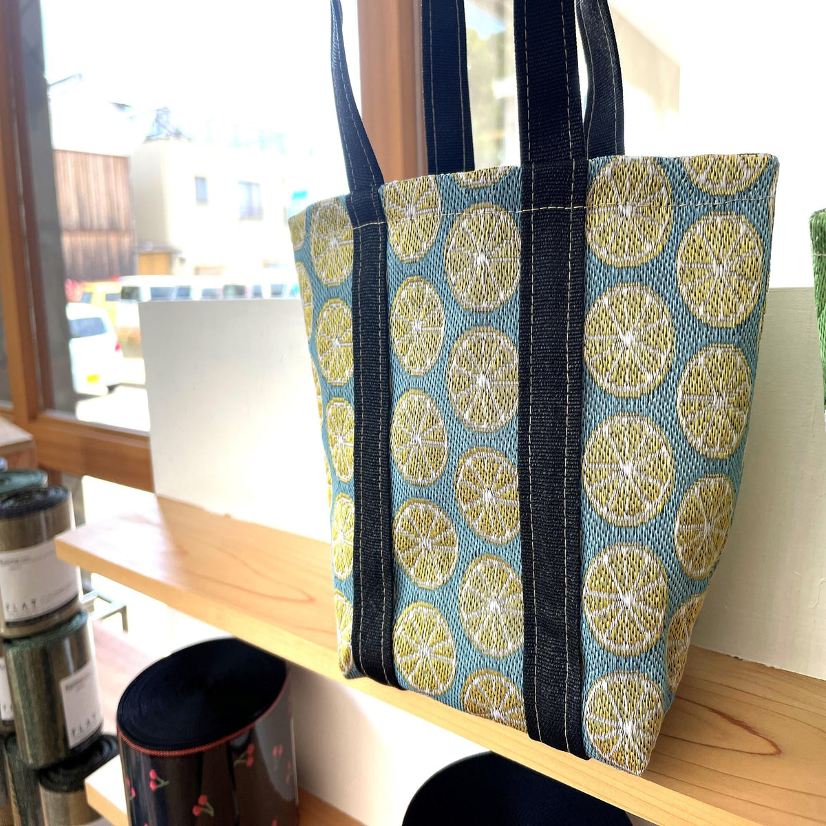 A handbag with a vibrant lemon pattern is displayed on a wooden shelf, with other items visible in the background.