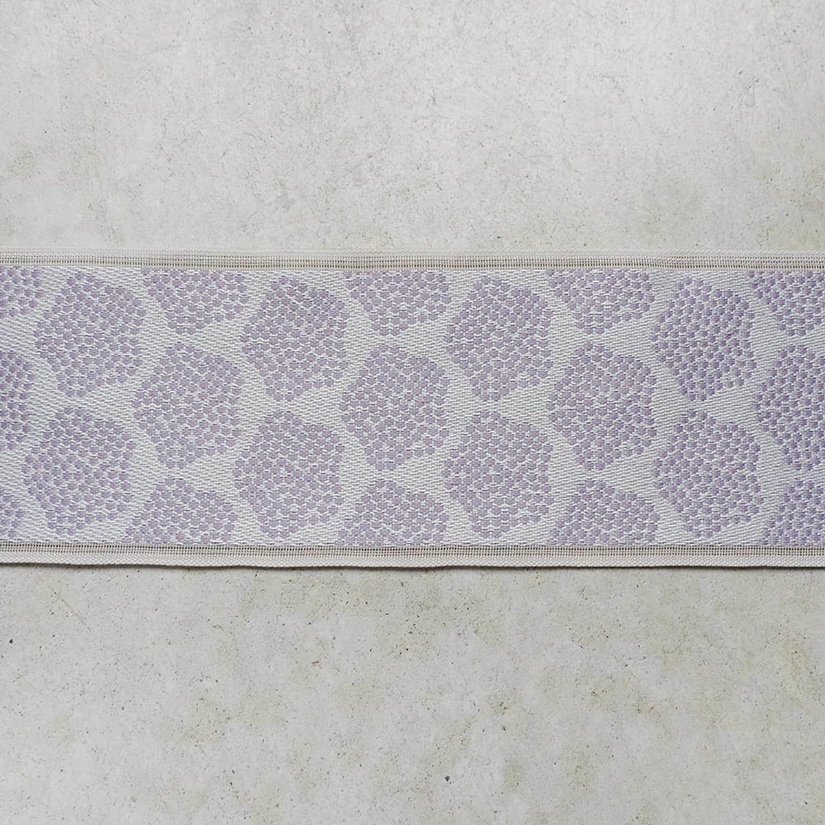 a close-up view of a fabric with a repeating pattern of purple flowers or leaves. The fabric appears to be a type of textile, possibly a piece of clothing or a decorative item. The pattern is intricate and detailed, with the flowers or leaves arranged in a repeating design across the fabric.