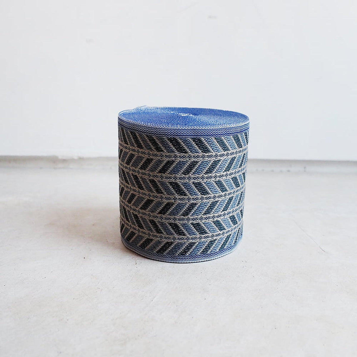 A blue and gray woven basket or container with a geometric pattern on its surface, sitting on a white surface against a plain background.