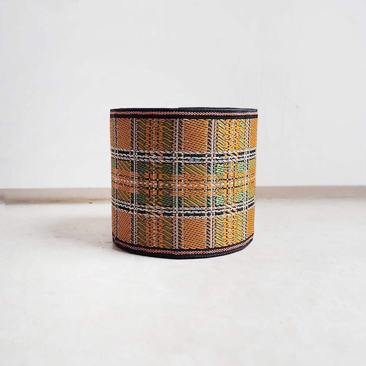 A cylindrical object with a plaid pattern in shades of orange, green, and black, sitting on a white surface against a plain background.