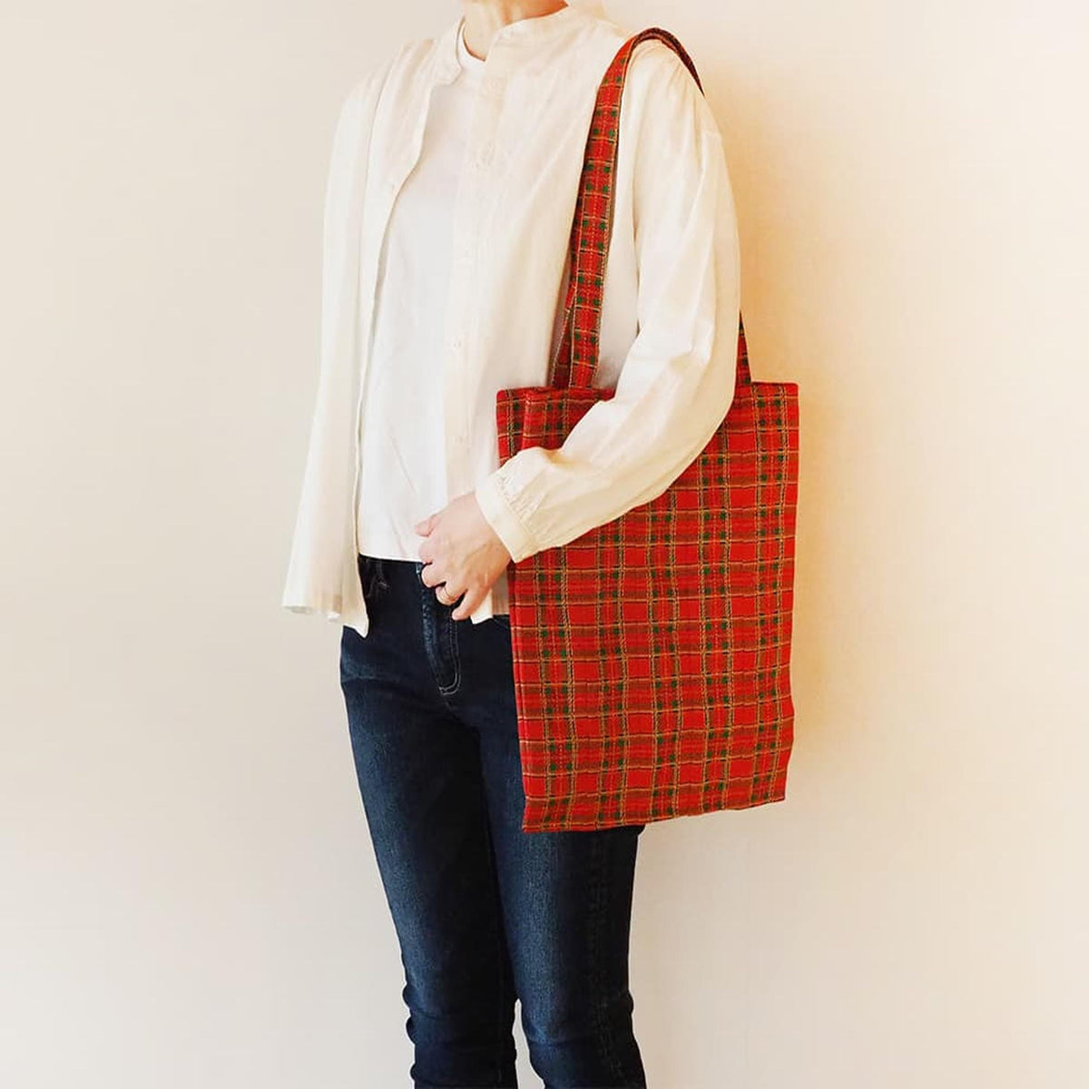 A person wearing a white blouse and blue jeans is holding a red and green plaid tote bag.