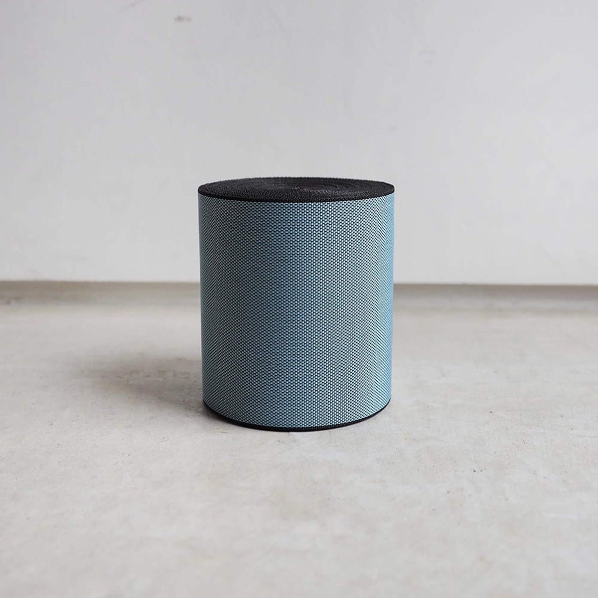A blue cylindrical object with a black top sits on a gray floor against a white wall.