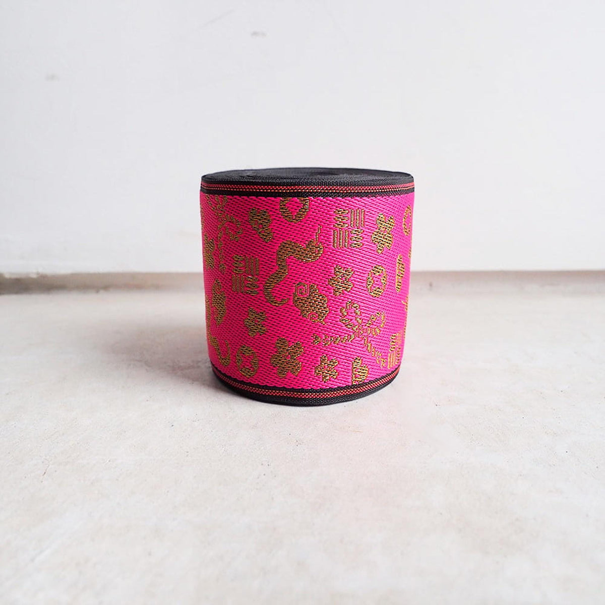 A pink and black woven container with a gold pattern on it, sitting on a white surface against a plain background.