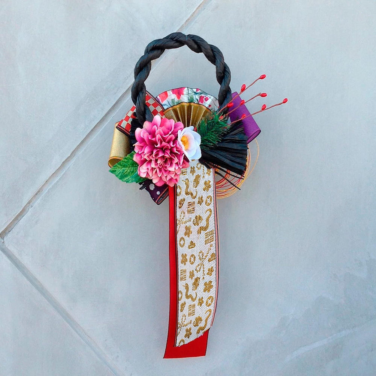 A decorative wreath made of various materials, including a braided handle, flowers, and a patterned ribbon, is displayed against a light gray background.