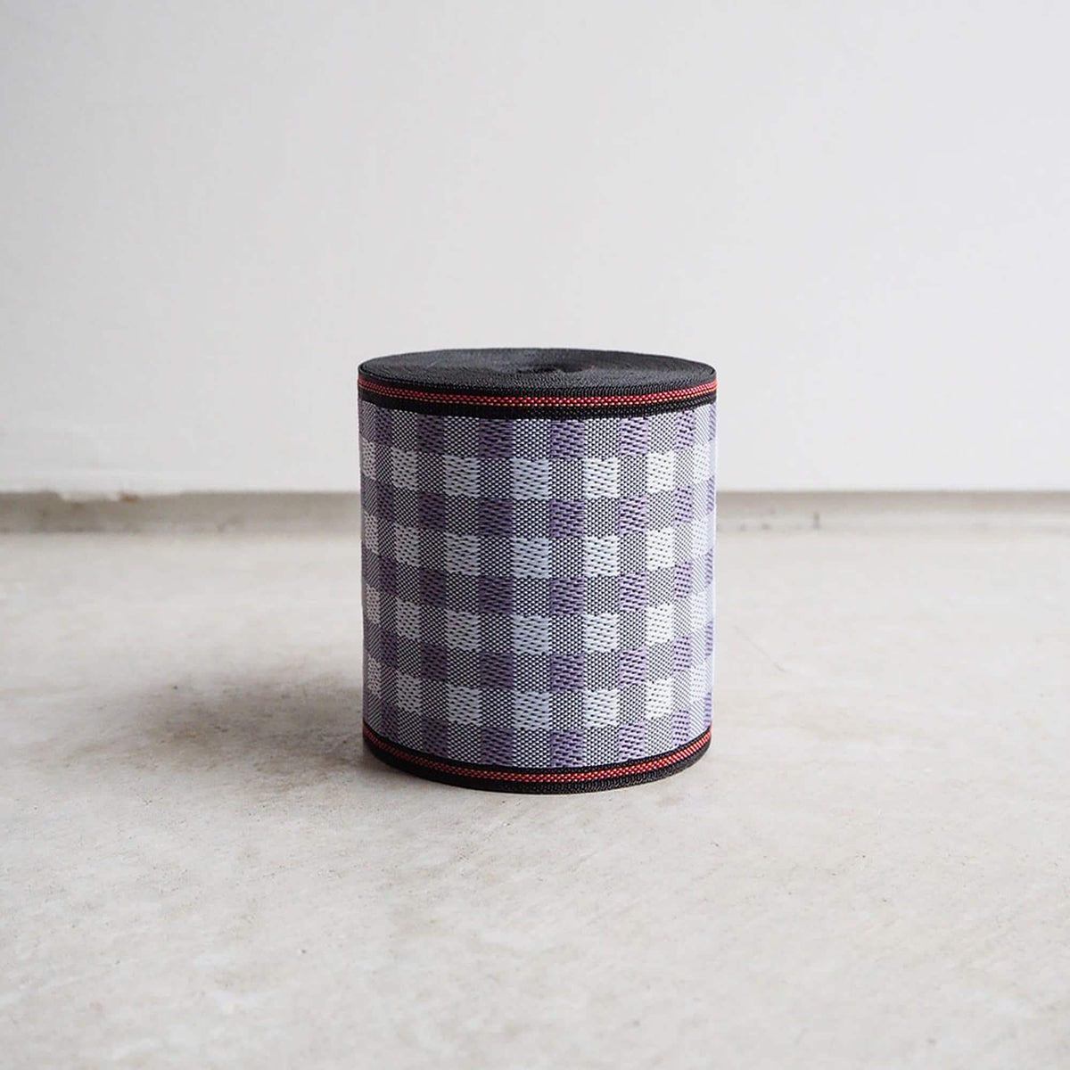 A cylindrical object with a checkered pattern in shades of purple and white, featuring a black border and red trim, is sitting on a plain surface.