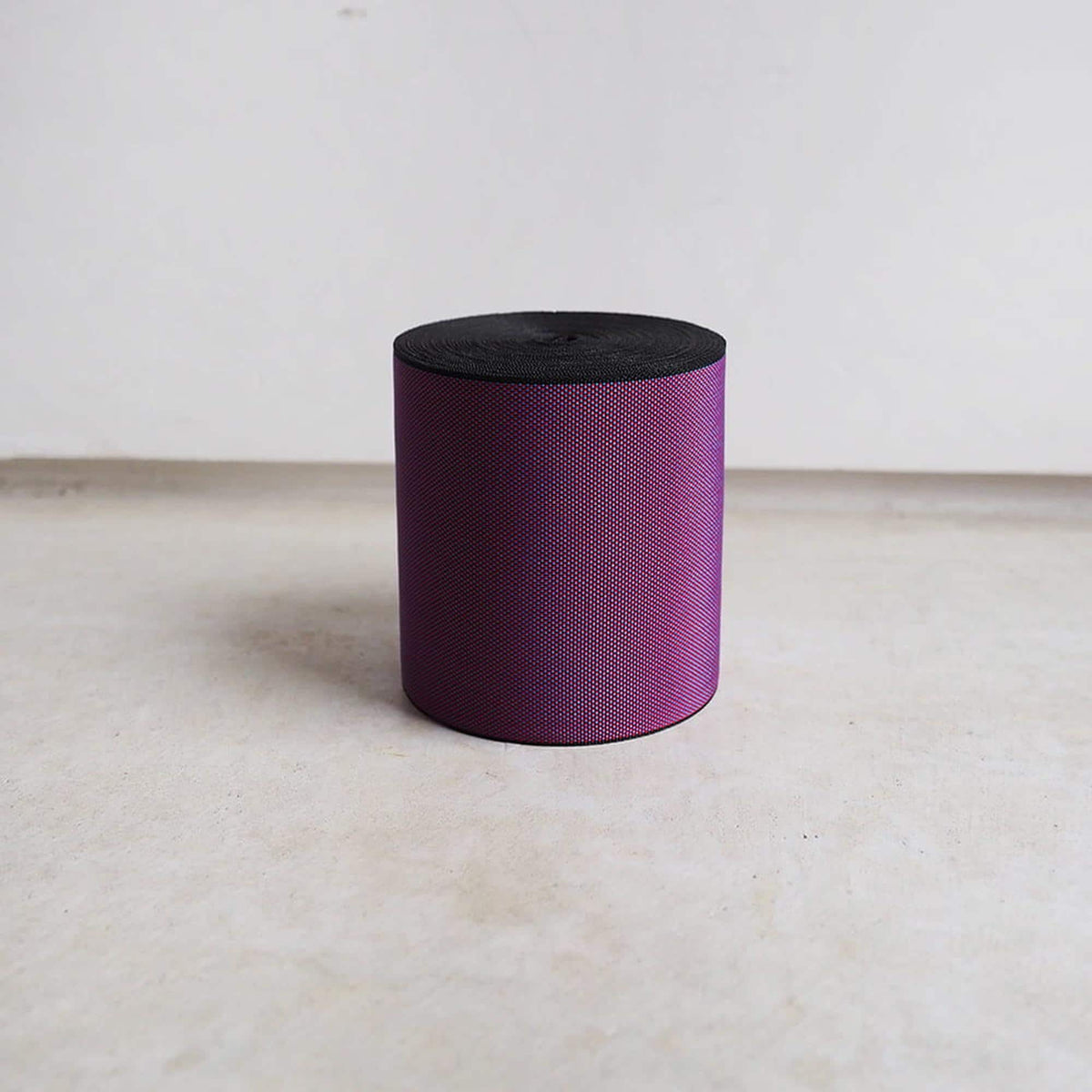 A purple cylindrical object with a black top is standing alone on a white surface against a plain white wall.
