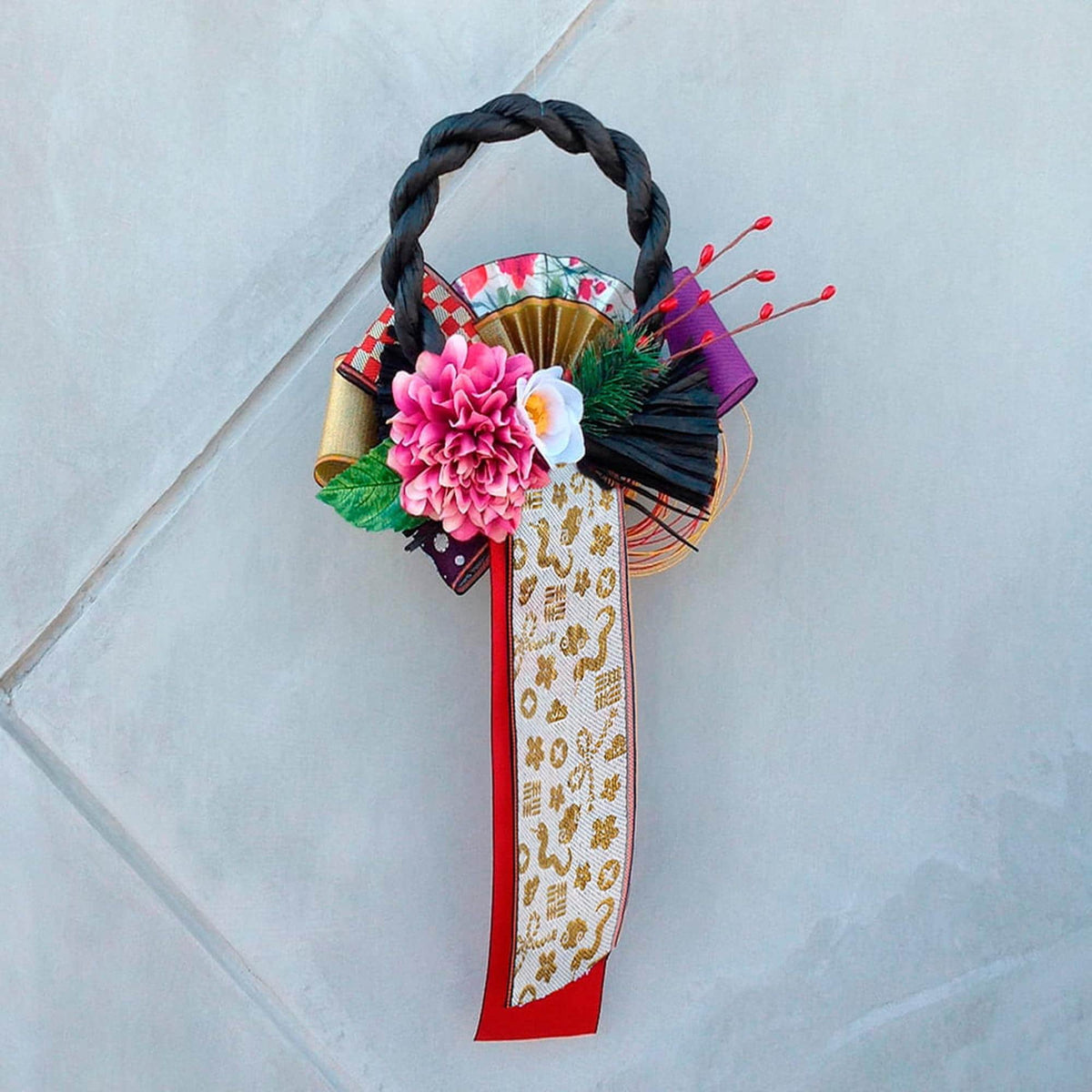 A decorative wreath made of various materials, including a braided handle, flowers, and a patterned ribbon, is displayed against a light gray background.