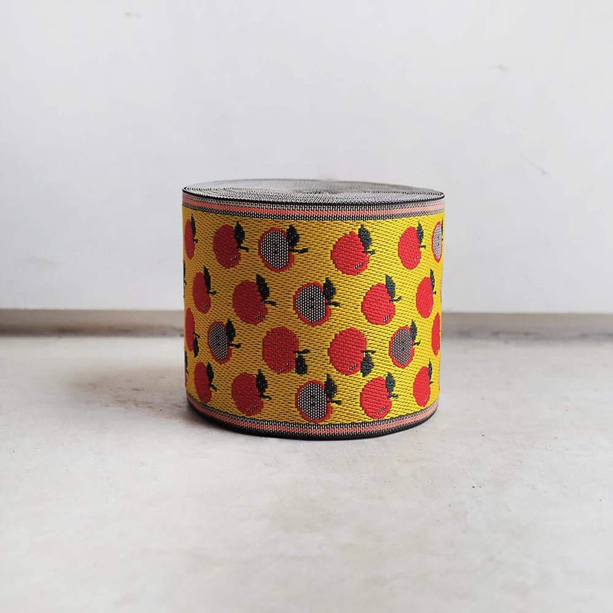 A round, cylindrical object with a pattern of red and yellow apples on it, sitting on a white surface against a plain background.