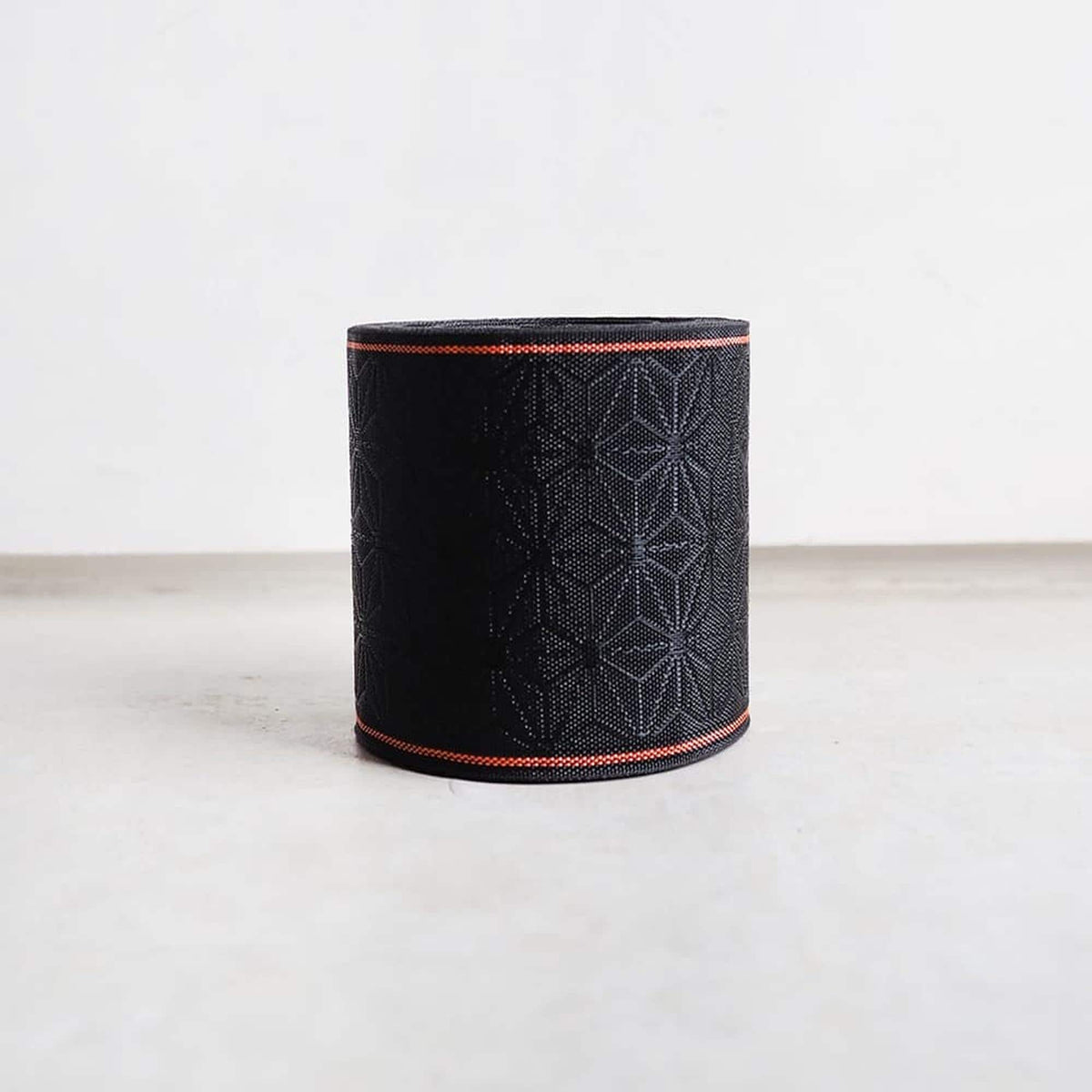A black cylindrical object with a red border, featuring a geometric pattern, is placed on a white surface against a plain white background.