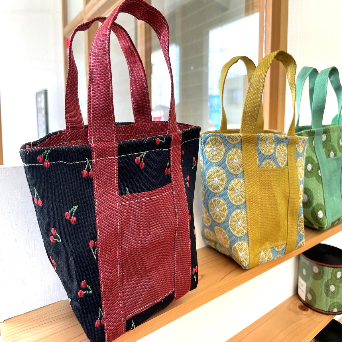 A row of colorful tote bags with various patterns, including red, black, yellow, and green designs, displayed on a wooden shelf.