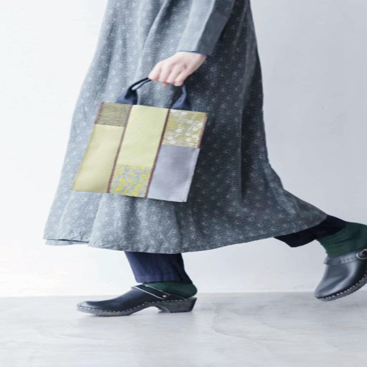 A person wearing a gray dress with a floral pattern and carrying a colorful bag is walking on a gray floor.