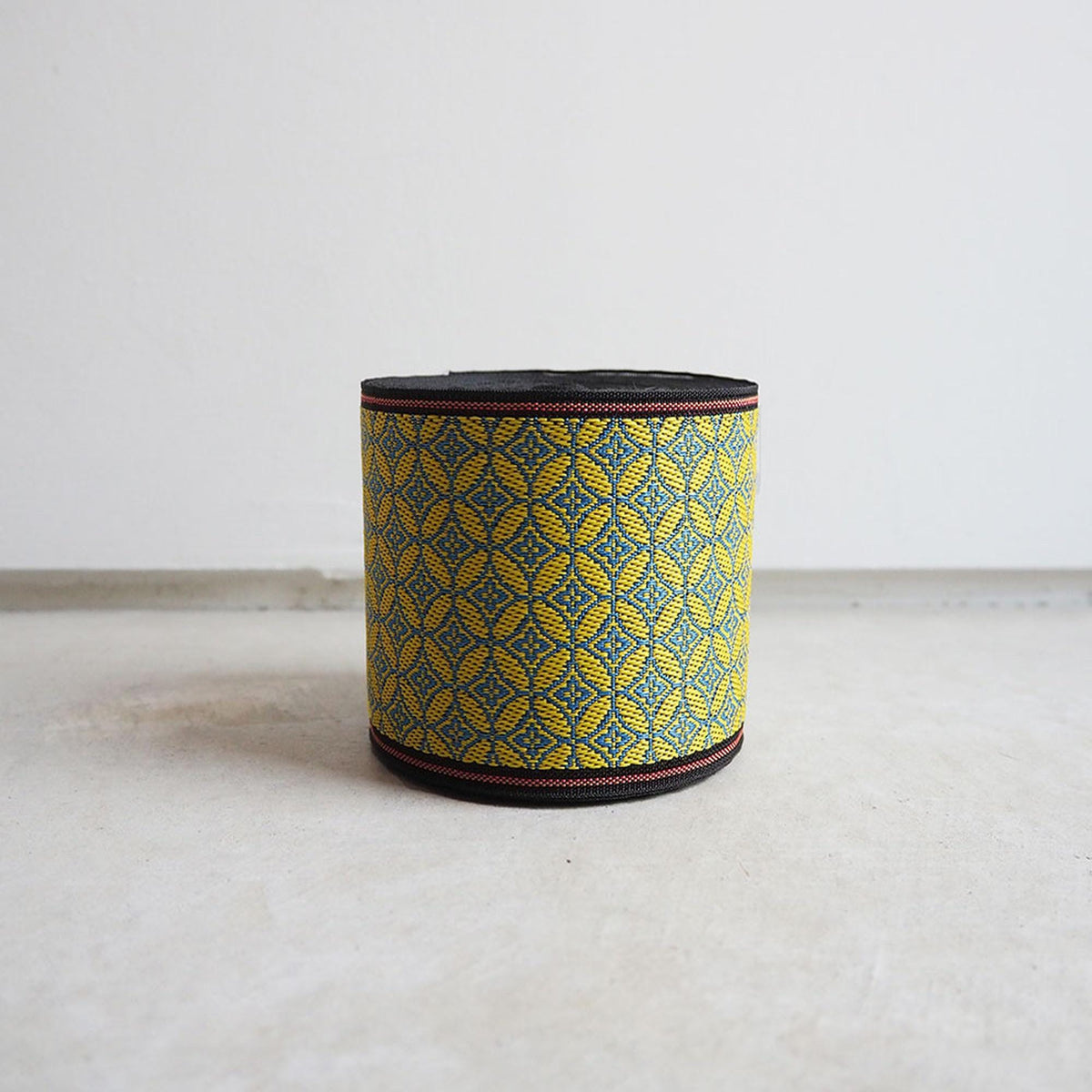 A cylindrical object with a black and yellow woven pattern, sitting on a white surface against a plain background.