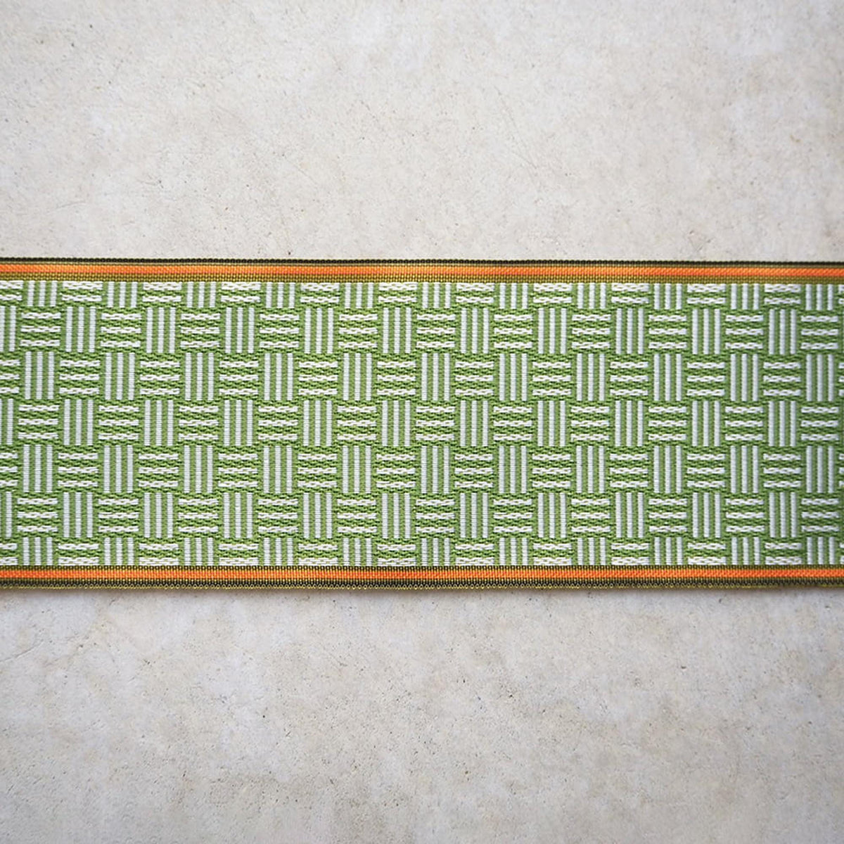 a close-up view of a woven fabric with a repeating pattern of green and white stripes. The fabric has a textured appearance, and the border is a combination of green and orange colors.