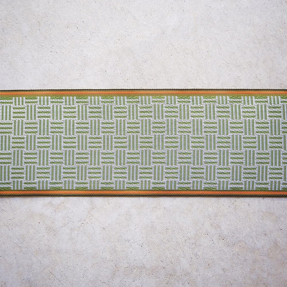 a close-up view of a woven fabric with a repeating pattern of green and white stripes. The fabric is placed on a plain, light-colored surface, such as a white wall or a concrete floor.