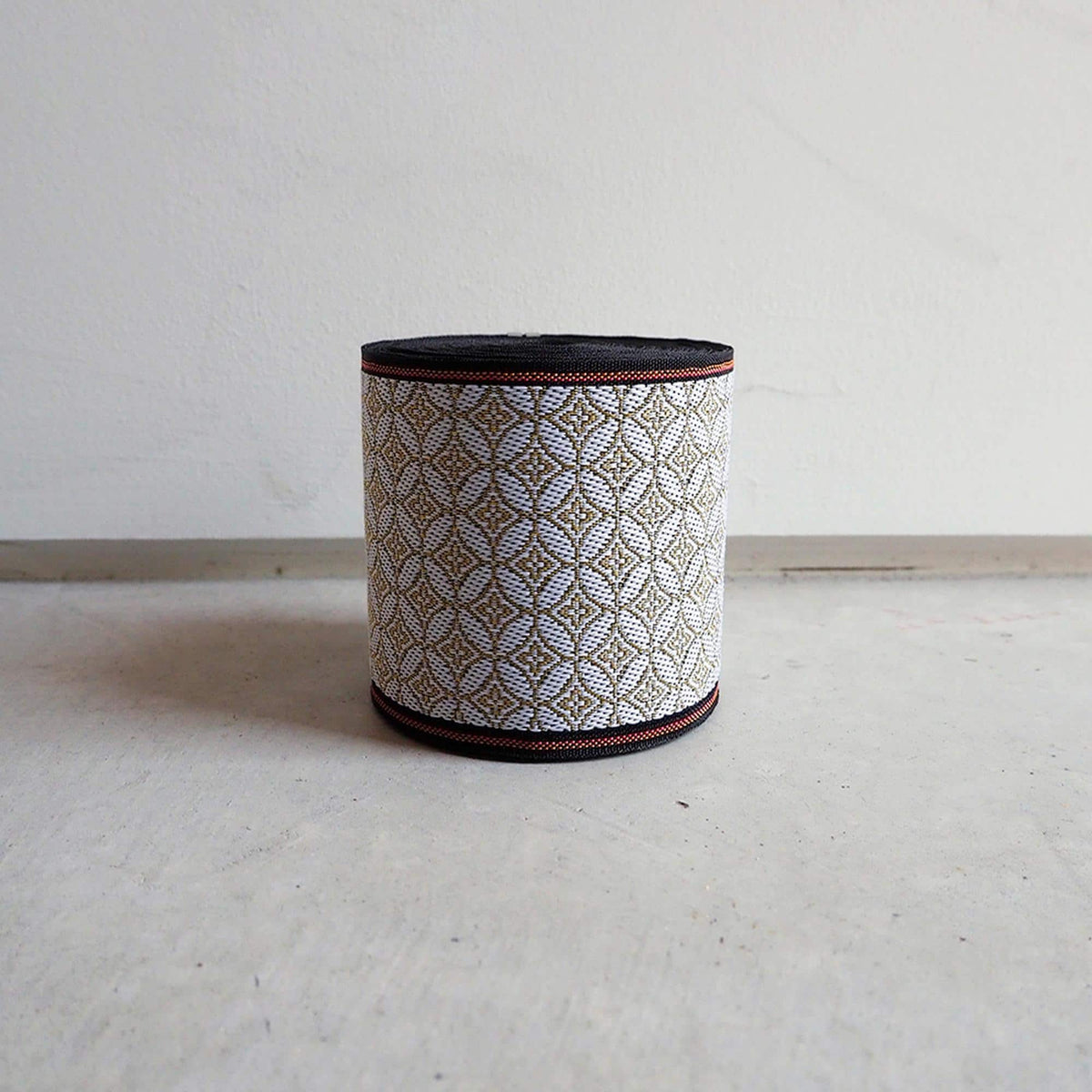 A cylindrical object with a patterned design on its surface, placed on a white surface against a plain white wall.