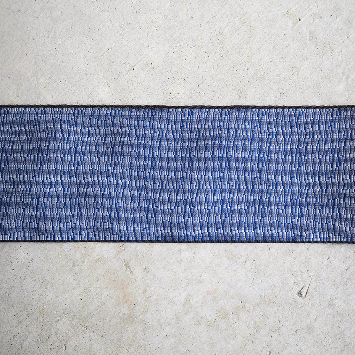 a close-up view of a blue and white patterned fabric or textile, which appears to be a rug or carpet. The fabric has a textured, woven appearance, and it is placed on a concrete surface.
