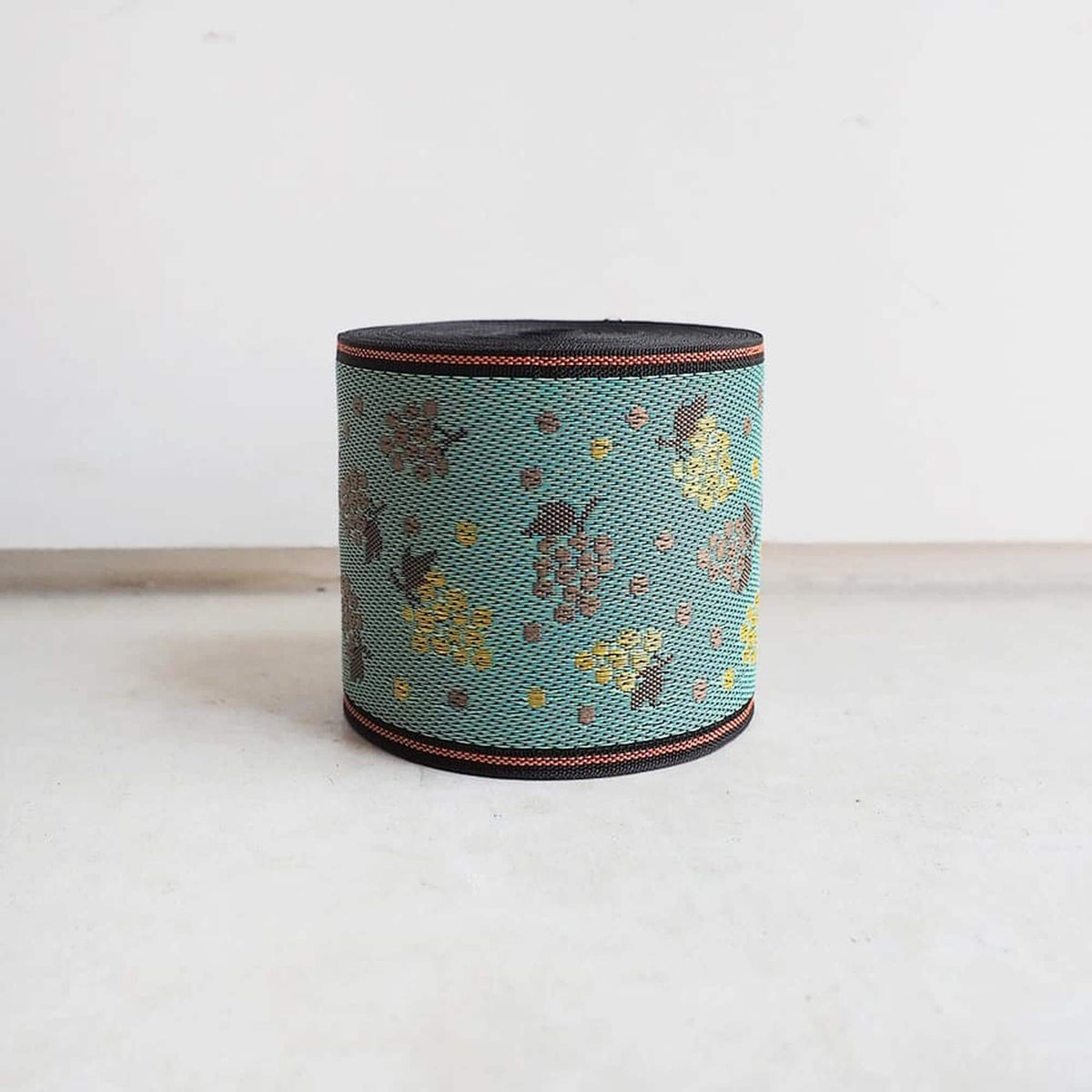 A cylindrical object with a pattern of small flowers or leaves on a turquoise background, sitting on a white surface against a plain white background.